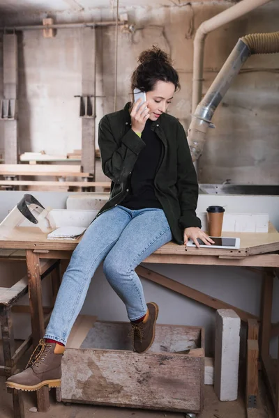 Woodworker talking on smartphone near digital tablet and coffee in workshop — Stock Photo