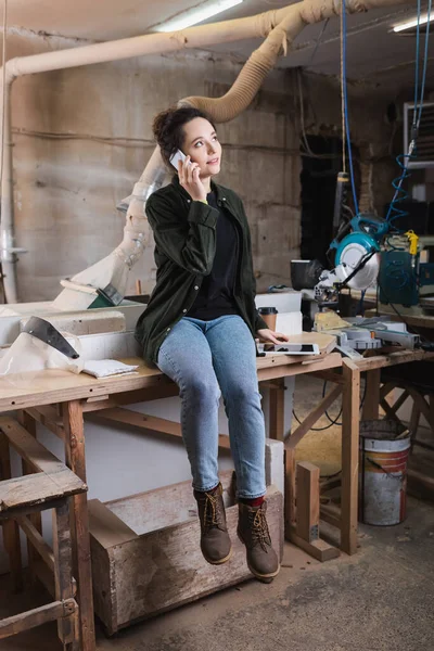 Young woodworker talking on smartphone near visor, coffee and digital tablet in workshop — Stock Photo