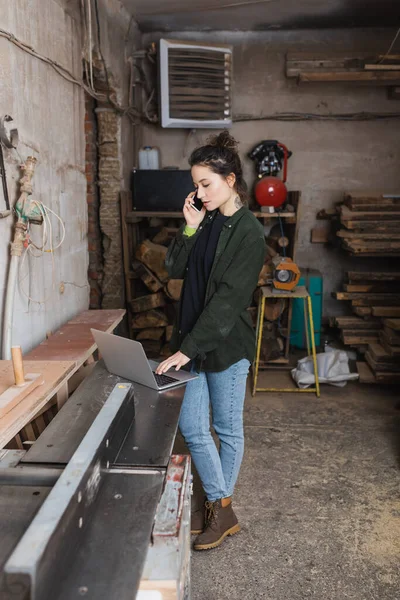 Brunette menuisier parler sur smartphone et en utilisant un ordinateur portable en atelier — Photo de stock