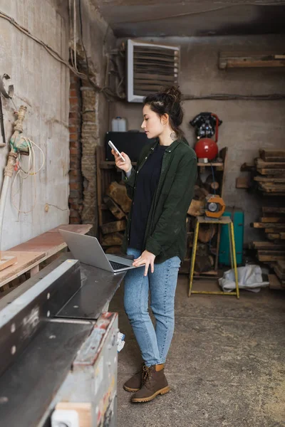 Vue latérale du menuisier à l'aide d'un téléphone portable près d'un ordinateur portable en atelier — Photo de stock