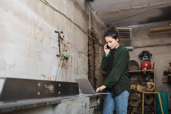 Tischler telefoniert mit Handy und Laptop in verschwommener Werkstatt — Stockfoto