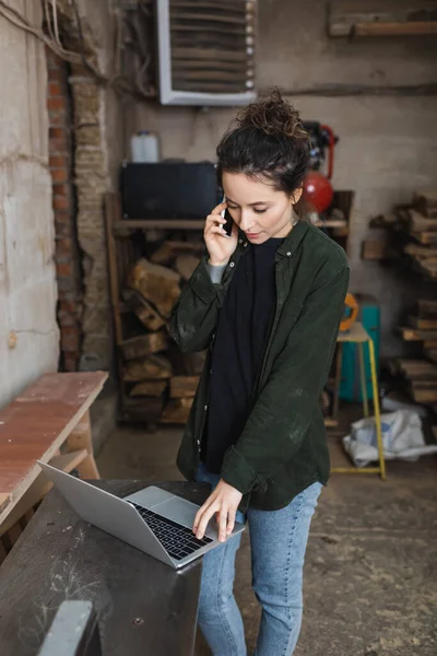 Morena woodworker falando no smartphone e usando laptop na oficina — Fotografia de Stock