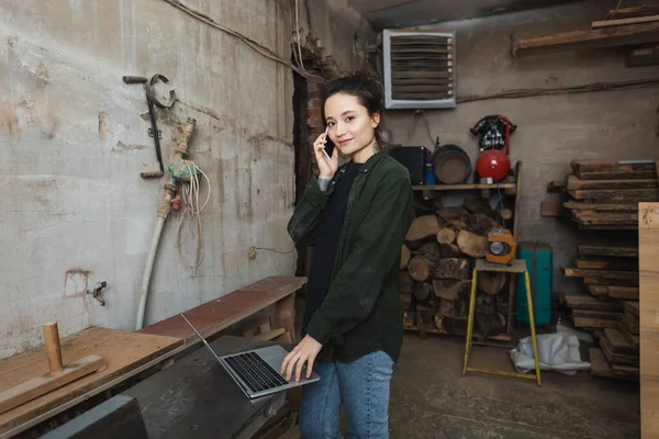 Smiling carpenter looking at camera while talking on smartphone and using laptop in workshop — Stock Photo