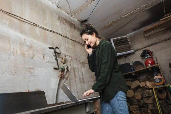 Positive carpenter using laptop and talking on cellphone in workshop — Stock Photo