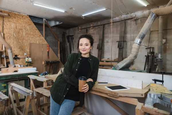 Fröhlicher Tischler hält Coffee to go in der Werkstatt und blickt in die Kamera — Stockfoto