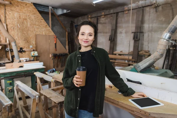 Smiling carpenter holding paper cup near devices in workshop — Stock Photo