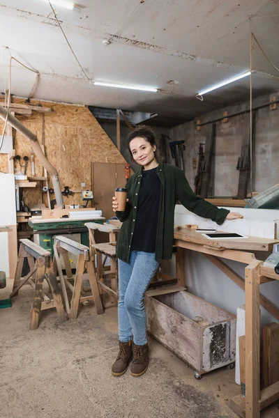 Joyeux concepteur de meubles tenant tasse en papier près des gadgets et de l'équipement dans l'atelier — Photo de stock