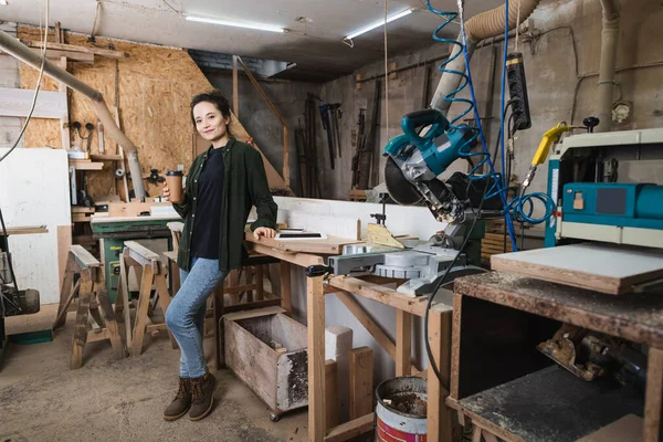 Diseñador de muebles sonrientes sosteniendo café para ir cerca de gadgets y equipos en el taller - foto de stock