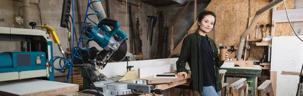 Joyeux menuisier tenant tasse en papier près des appareils avec écran blanc dans l'atelier, bannière — Photo de stock