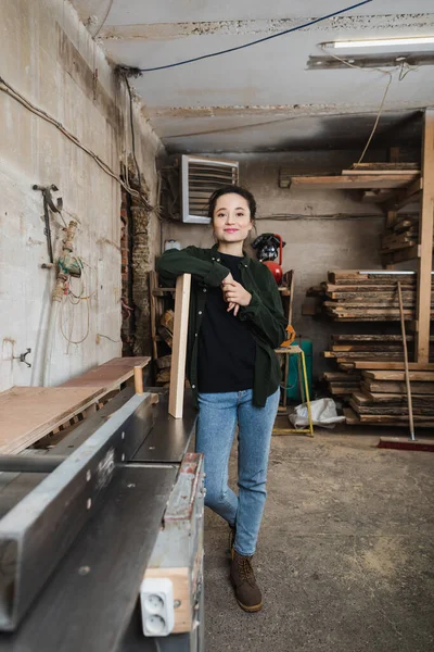 Joyeux menuisier regardant la caméra près de la planche et de la menuiserie — Photo de stock