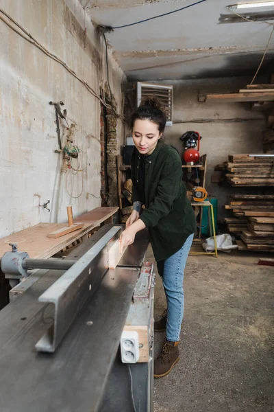 Designer de meubles tatoués en chemise tenant la planche près de la machine de menuiserie dans l'atelier — Photo de stock