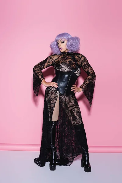 Vue pleine longueur de l'homme en perruque violette, robe en dentelle noire et corset en cuir debout avec les mains sur les hanches sur rose — Photo de stock