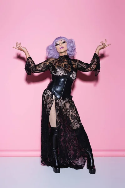 Exzentrische Drag Queen in schwarzem Spitzenkleid, lila Perücke und Lederstiefeln posiert auf rosa — Stockfoto