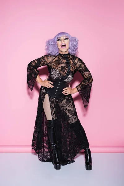 Astonished drag queen in violet wig and black lace dress looking at camera on pink — Stock Photo