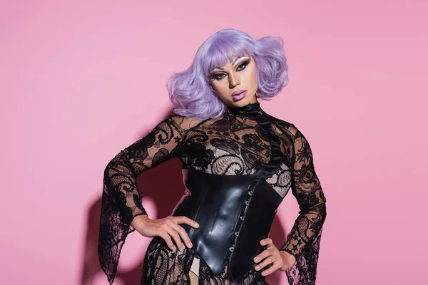 Man in purple wig, black lace dress and leather corset posing with hands on hips on pink — Stock Photo