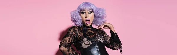 Astonished drag queen in black lace clothing and violet wig looking at camera on pink, banner — Stock Photo