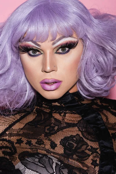 Retrato de drag queen en ropa de encaje negro y peluca púrpura mirando a la cámara aislada en rosa - foto de stock