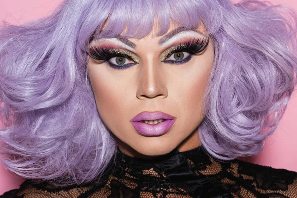 Retrato de hombre con maquillaje brillante, vistiendo peluca púrpura y mirando a la cámara en rosa - foto de stock