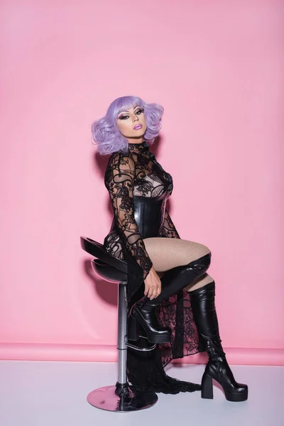 Glamour drag queen in violet wig and dress looking at camera while sitting on pink — Stock Photo