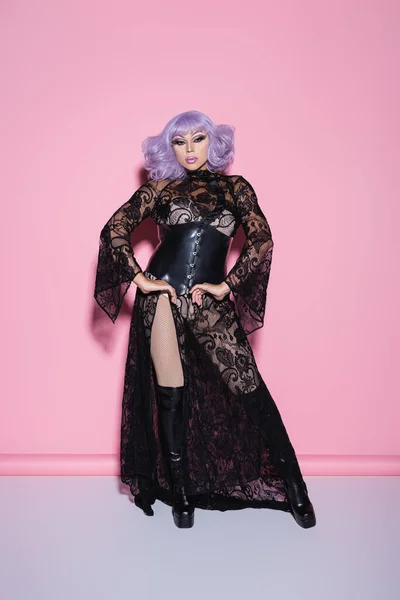 Vista completa de glamour drag queen en peluca púrpura y vestido de encaje de pie con las manos en las caderas en rosa - foto de stock