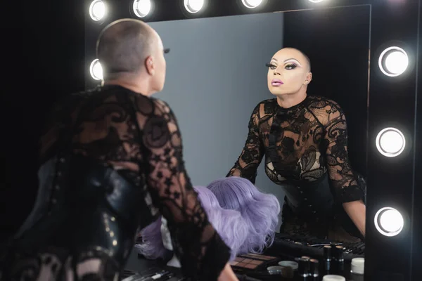 Homme dans un maquillage lumineux et des vêtements en dentelle noire regardant dans le miroir dans le vestiaire — Photo de stock