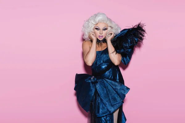 Glamour drag queen en vestido azul posando con las manos cerca de la cara mientras mira la cámara en rosa - foto de stock