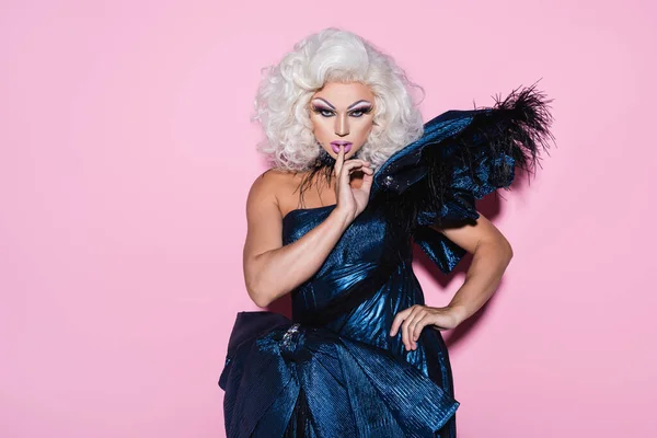 Drag queen en vestido azul mostrando signo de silencio mientras está de pie con la mano en la cadera en rosa - foto de stock