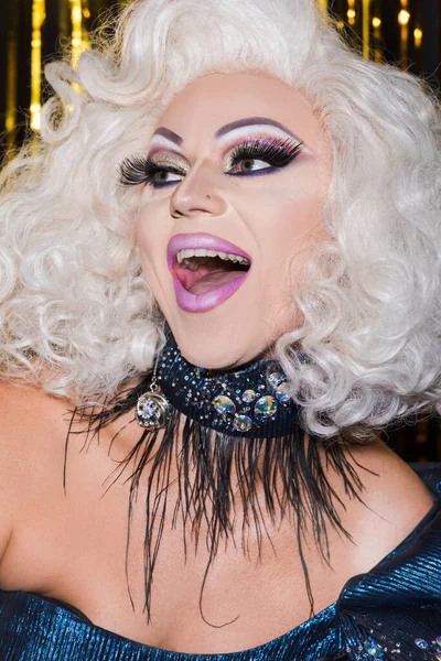 Возбужденный трансвестит с впечатляющим макияжем и открытым ртом на блестящем фоне — стоковое фото