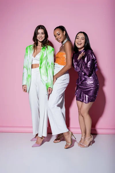 Full length of cheerful woman smiling with young interracial friends in stylish outfits on pink — Stock Photo