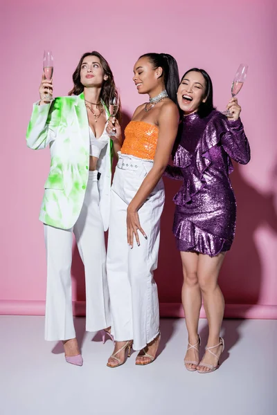 Comprimento total de mulheres inter-raciais alegres em roupas da moda segurando copos de champanhe em rosa — Fotografia de Stock