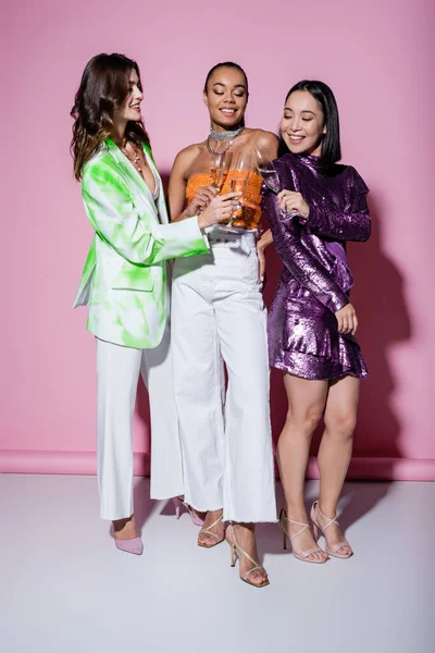 Comprimento total de mulheres inter-raciais alegres em roupas da moda copos clinking de champanhe em rosa — Fotografia de Stock