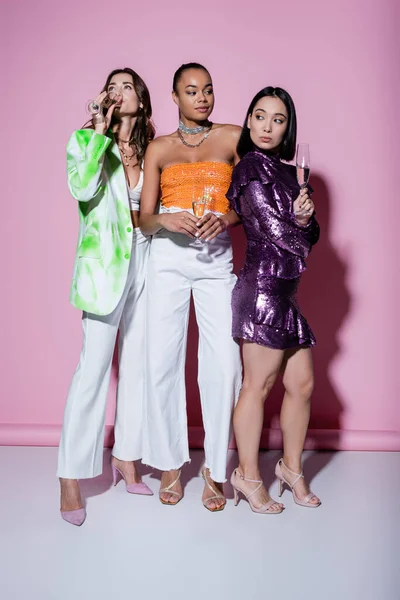 Longitud completa de las mujeres multiétnicas en trajes de moda de pie cerca de amigo beber champán en rosa — Stock Photo