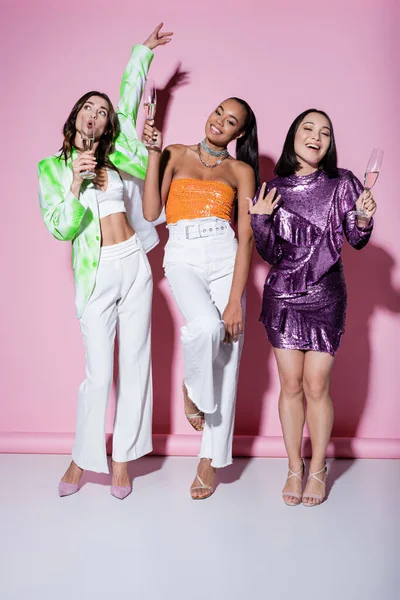 Comprimento total de mulheres multiétnicas alegres em roupas da moda segurando copos de champanhe durante a festa em rosa — Fotografia de Stock