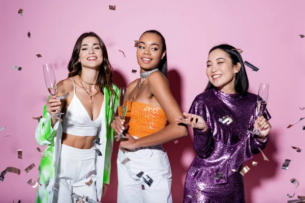 Alegres mujeres multiétnicas en trajes de moda sosteniendo copas de champán cerca de caer confeti en rosa - foto de stock