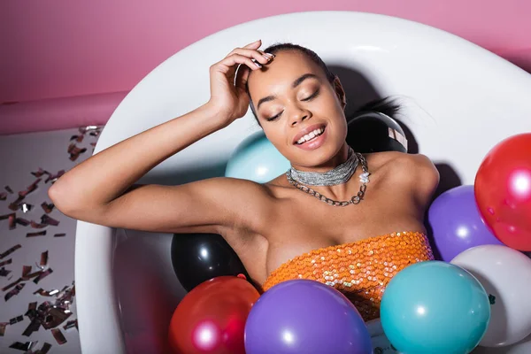 Glückliche Afroamerikanerin liegt in Badewanne mit bunten Luftballons auf rosa — Stockfoto