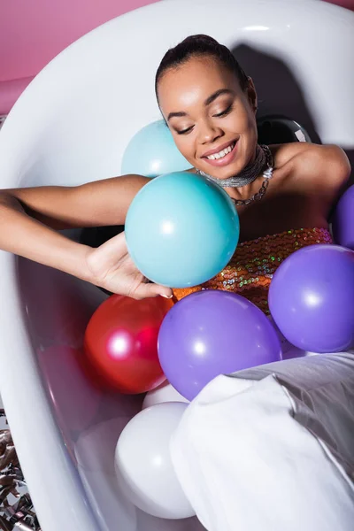 Високий кут зору щасливої афроамериканки лежить у ванні з різнокольоровими повітряними кулями на рожевому — стокове фото