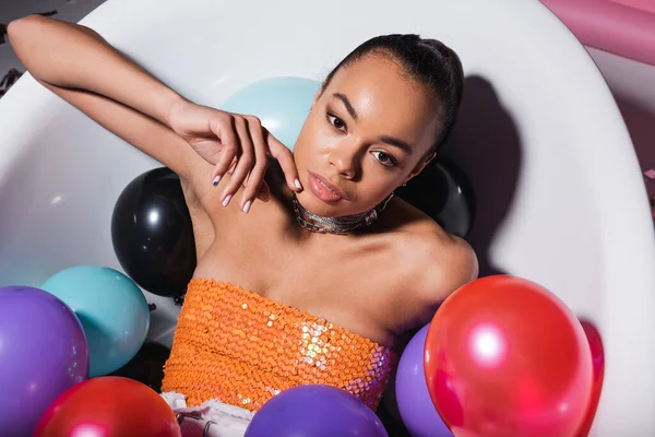 Vue grand angle de la jeune femme afro-américaine couchée dans la baignoire avec des ballons colorés — Photo de stock