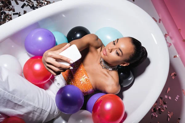 High angle view of african american model lying in bathtub with colorful balloons and taking selfie on smartphone — Stock Photo