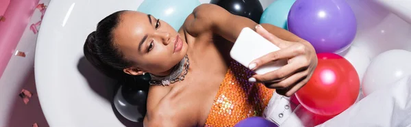 High angle view of african american model lying in bathtub with colorful balloons and taking selfie on smartphone, banner — Stock Photo