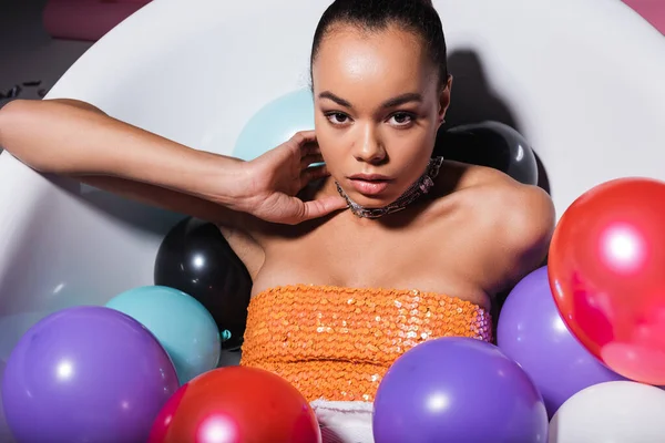 Happy african american woman lying in bathtub with colorful balloons looking at camera — Stock Photo