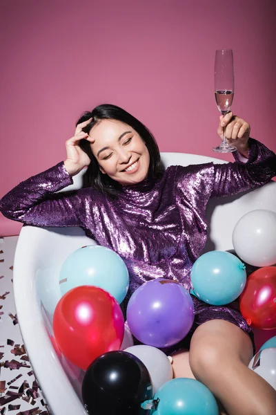 Happy asian woman in purple dress lying in bathtub with colorful balloons and holding glass of champagne on pink — Stock Photo