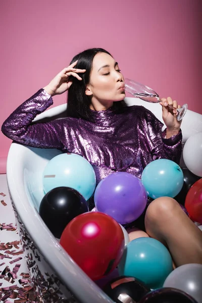 Asian woman in purple dress lying in bathtub with colorful balloons and drinking champagne on pink — Stock Photo