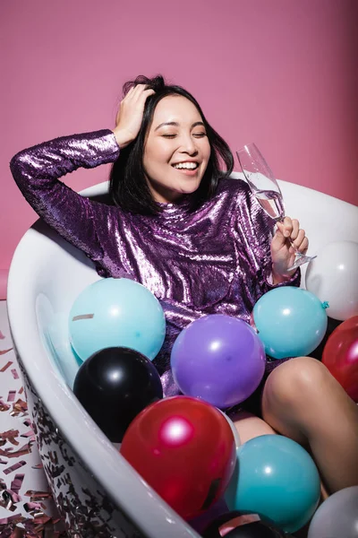 Cheerful asian woman in purple dress lying in bathtub with colorful balloons and holding glass of champagne on pink — Stock Photo