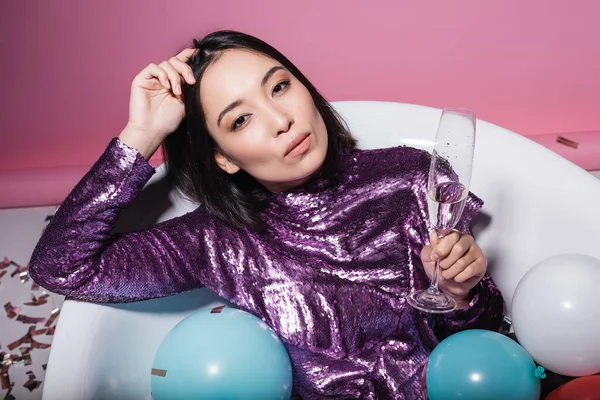 Jeune femme asiatique en robe pourpre couché dans la baignoire avec des ballons et tenant un verre de champagne sur rose — Photo de stock