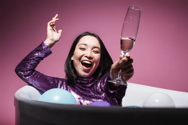 Excitada mujer asiática en vestido púrpura acostada en bañera con globos y sosteniendo copa de champán en rosa - foto de stock