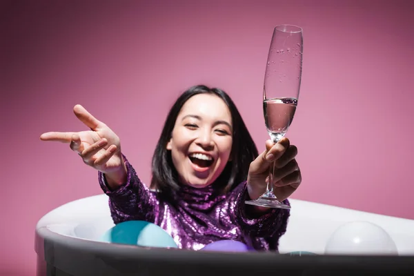 Amazed asian woman in purple dress lying in bathtub with balloons and holding glass of champagne on pink — Stock Photo