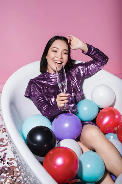 Здивована азіатська жінка в одязі лежить у ванні з різнокольоровими повітряними кулями і тримає келих шампанського на рожевому — стокове фото