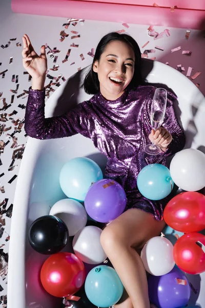 Vue grand angle de femme asiatique heureuse couchée dans la baignoire avec des ballons colorés et tenant un verre de champagne — Photo de stock