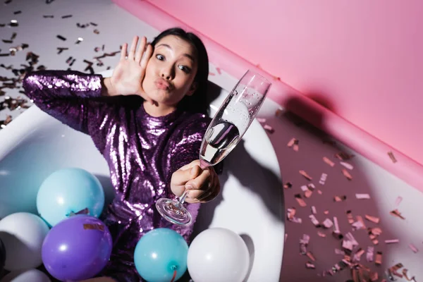 Vue grand angle de femme asiatique boudant lèvres tout en étant couché dans la baignoire avec des ballons et tenant verre flou de champagne — Photo de stock