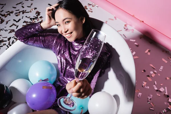 Vue grand angle de femme asiatique heureuse couchée dans la baignoire avec des ballons et tenant un verre de champagne près de confettis — Photo de stock